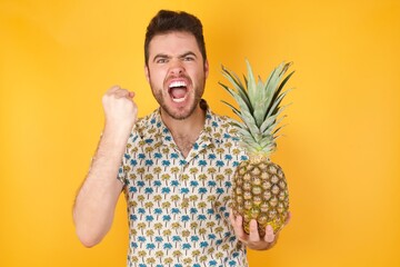 Young man holding pineapple wearing hawaiian shirt over yellow isolated background  being angry and...