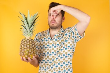 Young man holding pineapple wearing hawaiian shirt over yellow isolated background  smelling...