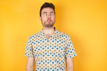 Young man holding pineapple wearing hawaiian shirt over yellow isolated background  making fish face with lips, crazy and comical gesture. Funny expression.