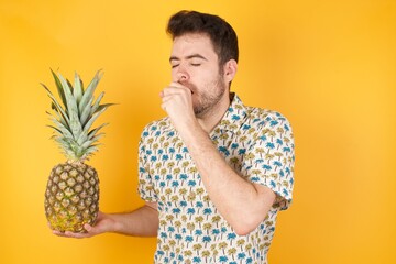 Young man holding pineapple wearing hawaiian shirt over yellow isolated background  feeling unwell and coughing as symptom for cold or bronchitis. Healthcare concept.