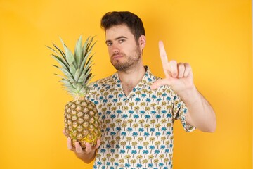 Young man holding pineapple wearing hawaiian shirt over yellow isolated background  making fun of...