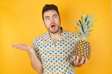 Frustrated Young man holding pineapple wearing hawaiian shirt over yellow isolated feels puzzled...