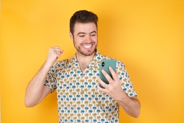 Portrait of Young man holding pineapple wearing hawaiian shirt over yellow isolated background holding in hands cell reading browsing 