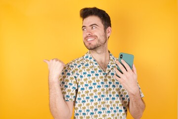 Young man holding pineapple wearing hawaiian shirt over yellow isolated background using and texting with smartphone pointing and showing with thumb up to the side with happy face smiling