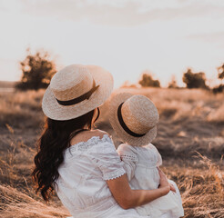 brunette mother and little daughter are sitting on the dry grass in white dresses. On the Sunset....