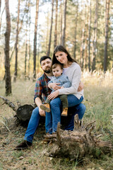 Naklejka na ściany i meble Outdoor portrait of happy stylish Caucasian family in casual clothes, young parents and cute little child son, on a walk in beautiful pine forest, sitting on a log of old tree and looking at camera