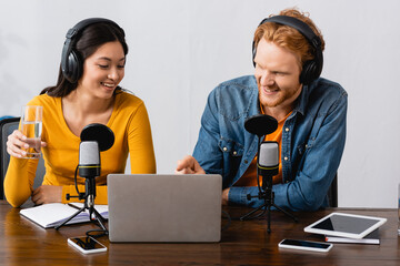 young announcer pointing with finger at laptop near asian colleague holding glass of water