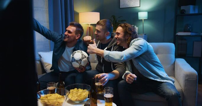 Three cheerful Caucasian friendly men sitting on sofa at night with football ball, snacks and beer and taking selfie photo with smartphone camera. Sport fans posing to phone in living room at TV.