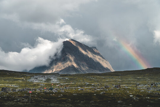 Dramatic view of a rugged mountain along Sweden&rsquo;s Kungsleden trail, with its rocky slopes lit by soft sunlight and partly wrapped in swirling clouds. In the foreground, a marked winter trail and a lon