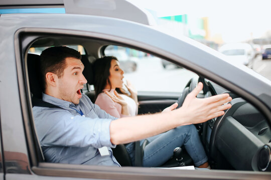 Male auto instructor takes exam in young woman. Scared frightened man point forward with both hands. Female stident driwing without steering wheel. Scared model screaming.