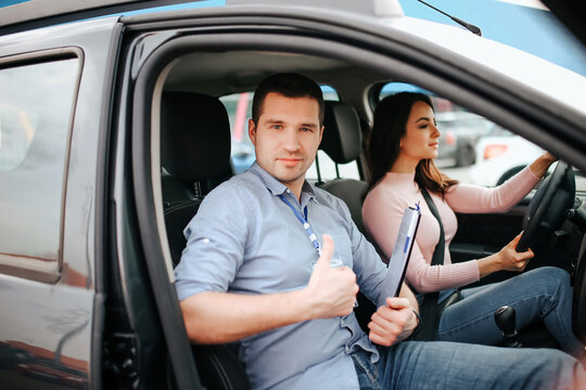 Male auto instructor takes exam in young woman. Happy positive young man look on camera and hold big thumb up. Careful young woman sriving car. Sit on driver's place and hold hands on steering wheel.