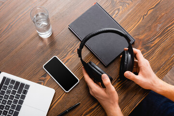 cropped view of freelancer holding wireless headphones near smartphone with blank screen, laptop, notebook and glass of water - obrazy, fototapety, plakaty