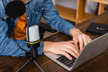 cropped view of broadcaster in denim shirt typing on laptop near microphone
