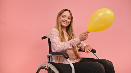 Young disabled woman in wheelchair holding yellow balloon, isolated on pink background. High...