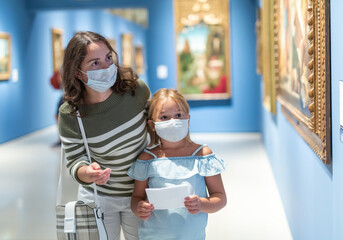 Portrait of woman with girl in medical masks standing in museum of art and looking at painting