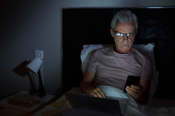 Senior man in eyeglasses lying on bed and using his mobile phone and laptop in online work he working till late