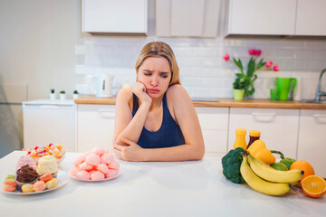 Obraz na płótnie Canvas Diet struggle. Young sad woman in blue T-shirt choosing between fresh fruit vegetables or sweets in the kitchen. Choice between healthy and unhealthy food. Dieting. Healthy Food