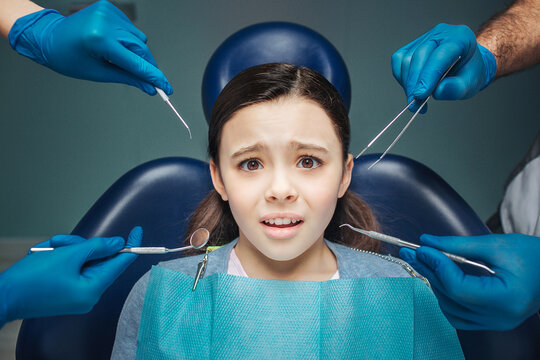 Scared girl sit in detal chair in room. She looks scared and frightened. Adult's hands in latex gloves hold dentist tools.