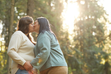 Two overweight lesbians kissing each other during their walk in the forest - obrazy, fototapety, plakaty