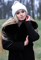 Blonde girl posing at the winter city in a black fur coat and a white hat