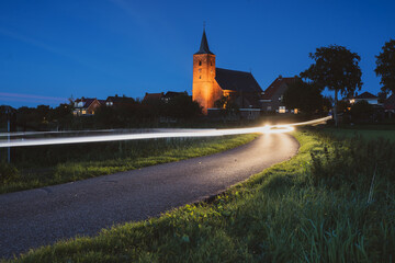 Obraz premium Car lighting streaks of light drive past a small village church in a Dutch landscape