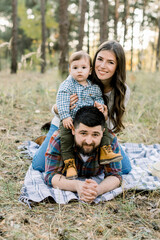 Family, people, love and happiness concept. Outdoor portrait of young attractive family, dad, mom and kid son, lying on the checkered blanket in autumn pine forest and smiling to camera