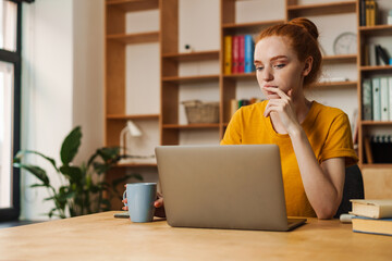 Image of serious redhead girl working with laptop while sitting at table