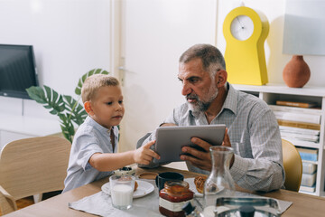 boy using tablet while having breakfast at home with his grandfather
