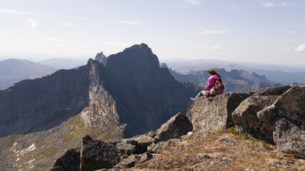The girl traveler sits on a high rock and admires the view of the mountains. Hiking in Ergaki. Backpack, windbreaker, boots