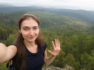 A girl traveler takes a selfie against a beautiful landscape. Smiling brunette. Hair pulled back in a ponytail, long hair. Sends greetings, hand raised. Beautiful green landscape. Hiking