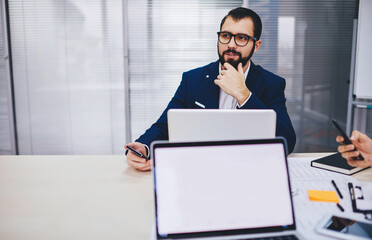 Thoughtful male employee in spectacles pondering on financial research during collaborative working process at desk with mockup laptop computer, blank netbook with copy space for business website