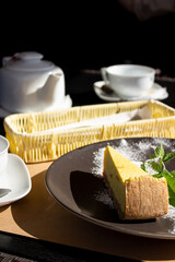 Close-up on a black table is a tea set with fruit tea, a black plate with cheesecake, mint and cutlery.