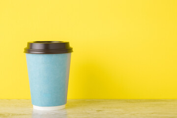 Blue disposable paper cup on wooden table at bright yellow wall. Takeaway coffee or other drink. Closeup. Empty place for text. Front view.