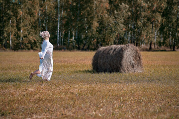 Naklejka na ściany i meble A girl in a white dress runs across the field to a haystack