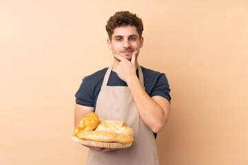 Male baker holding a table with several breads isolated on beige background laughing