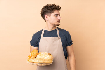 Male baker holding a table with several breads isolated on beige background looking side