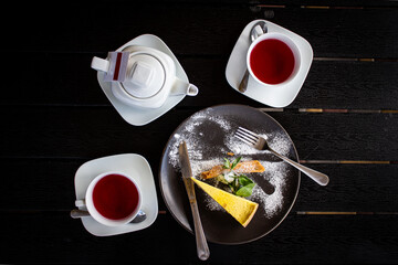 View from above. Close-up on a black table is a tea set with fruit tea, a black plate with cheesecake, mint and cutlery.