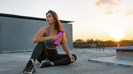 A sports girl with a metal bottle of water after training rests against the background of sunset