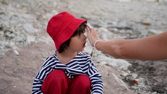 mom adjusts the hair of a boy child sitting on a stone beach in a red cap and vest