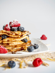Stack of pancakes with forest fruit, raspberry, blueberry, chocolate scirope and icing sugar on a light-colored plate. White background.