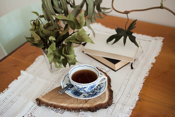 themed coffee corner with dried eucaliptus and bay leaves and retro tablecloth, autumn vibes, bohemian porcelain ornamental mug on the wooden cut with the bark, books in the background