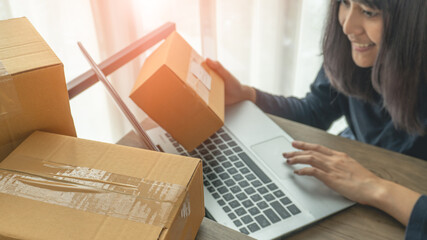 Smiling woman using a laptop for business at home on background and parcel boxes on desk in the foreground with copy space. Business start up SME concept.