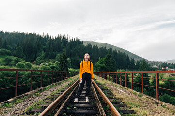 Stylish hipster woman in orange sweatshirt stands on the railway track against the backdrop of a mountain landscape and looks at the camera.