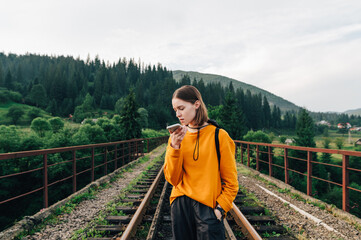 Fototapeta na wymiar Attractive female hiker in bright casual clothes stands in the mountains on the railway track and records audio messages on a smartphone. Vorokhta, Carpathians, Ukraine.