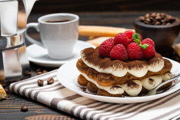 portion of Classic tiramisu dessert with raspberries, cup of espresso and coffee maker on wooden background