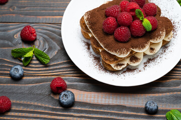 portion of Classic tiramisu dessert with raspberries and blueberries on wooden background