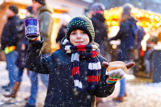 Little Cute Kid Boy Eating German Sausage And Drinking Hot Children Punch On Christmas Market. Happy Child On Traditional Family Market In Germany. Laughing Boy In Colorful Winter Clothes