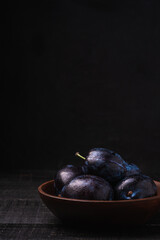 Fresh ripe plum fruits with water drops in wooden bowl on dark wood background, angle view