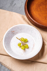 Fresh ripe white grape berries in wooden bowl with plate on linen tablecloth, stone concrete background, angle view