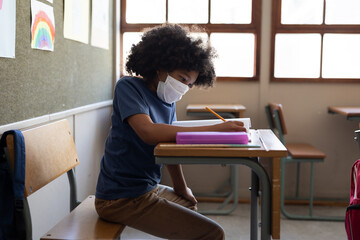 Boy wearing face mask writing while sitting on his desk at school